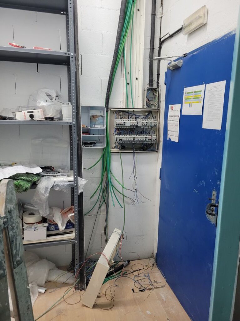 Área técnica o de almacenamiento con un cuadro eléctrico abierto mostrando cables y conexiones internas. Al lado hay una puerta azul con documentos pegados. A la izquierda, un estante metálico contiene varias herramientas, materiales y equipos desorganizados.