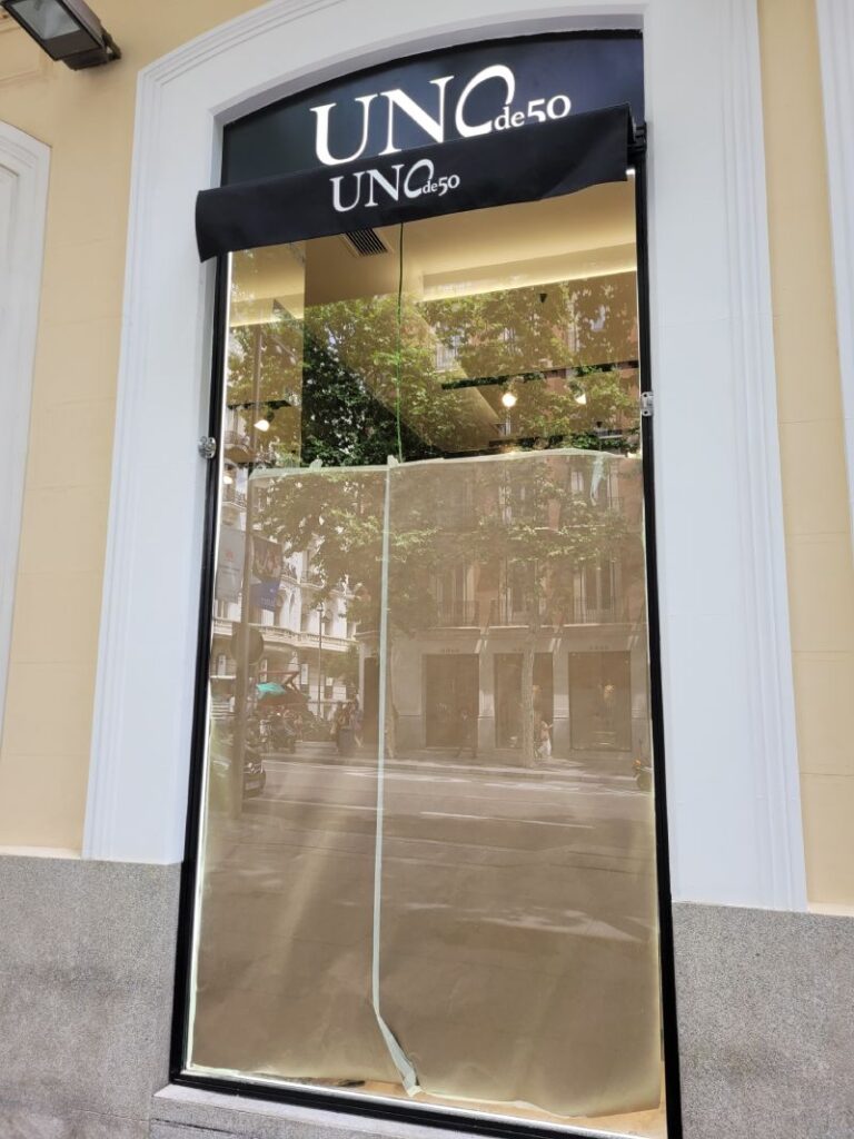 Vitrina de una tienda "UNO de 50" cubierta con papel kraft desde el interior. La tienda tiene un toldo negro con el nombre "UNO de 50" en letras blancas. La vitrina refleja la calle y los edificios cercanos, mostrando un entorno urbano con árboles y transeúntes. El marco de la vitrina es negro, y las paredes exteriores son de color claro con un zócalo de granito.