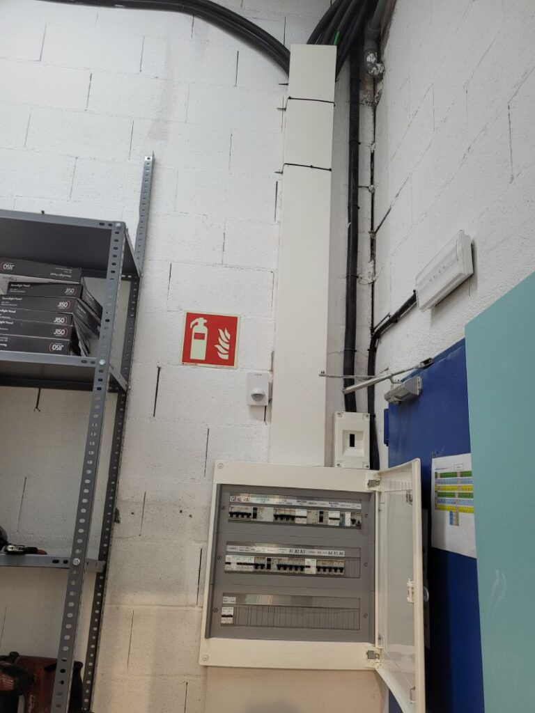 Área de almacenamiento o sala técnica con una caja de distribución eléctrica abierta, mostrando los interruptores y circuitos internos. La caja está montada en una pared de bloques de cemento pintados de blanco. Junto a la caja hay una puerta azul con documentos pegados y un estante metálico con materiales almacenados.