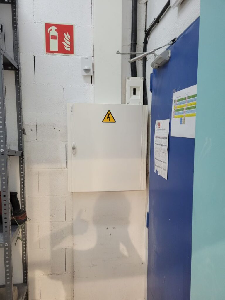 Área de almacenamiento o sala técnica con una caja de distribución eléctrica montada en la pared, señalizada con un símbolo de alto voltaje. Junto a la caja hay una puerta azul con documentos pegados y un estante metálico con herramientas y materiales. En la pared, hay un letrero de color rojo que indica la ubicación de un extintor de incendios. La pared es de bloques de cemento pintados de blanco, y hay cables y conductos visibles cerca del techo.