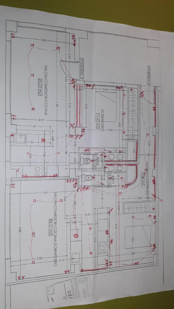 Plano arquitectónico de una vivienda que incluye anotaciones y marcas en color rojo. El plano muestra la distribución de diferentes áreas, como salón-comedor-cocina, dormitorio, aseo y vestíbulo distribuidor, con medidas y especificaciones detalladas. Las anotaciones en rojo parecen indicar modificaciones o instalaciones adicionales, como cableado eléctrico o tuberías.
