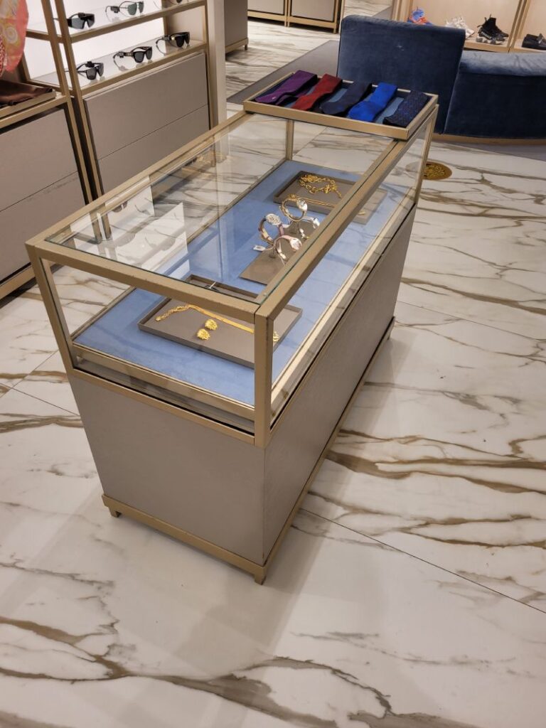 Vitrina de exhibición en una tienda de lujo, con un marco dorado y superficie de vidrio, mostrando joyería y accesorios sobre un fondo azul. La vitrina está colocada sobre un piso de mármol blanco con vetas grises. Al fondo, se pueden ver estanterías con gafas de sol y otros accesorios, así como un sofá azul y más productos exhibidos en la tienda. El ambiente es elegante y bien iluminado.