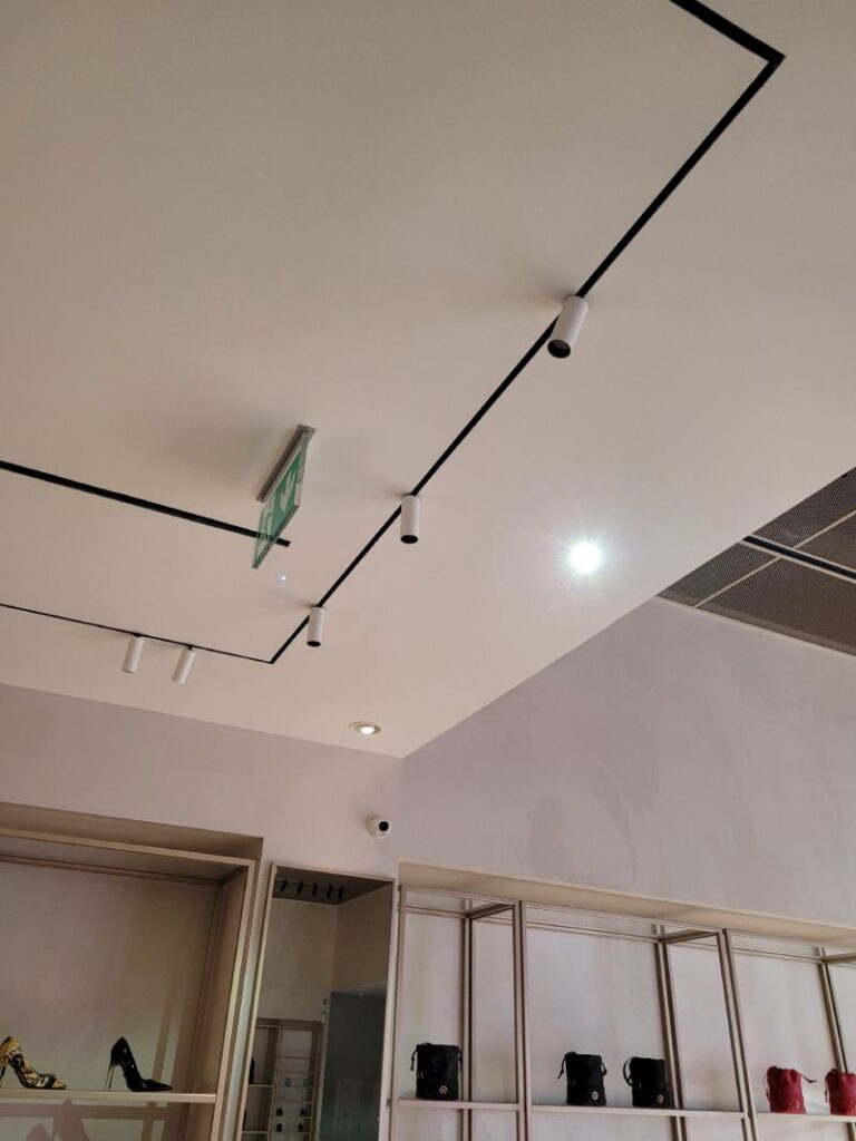 Techo de una tienda con rieles negros y focos LED cilíndricos blancos. Hay una señal de salida de emergencia verde colgando del techo. Abajo se ven estanterías de exhibición con zapatos y bolsos. El diseño interior es moderno y minimalista, con paredes de color claro y un enfoque en la iluminación y la presentación de los productos.