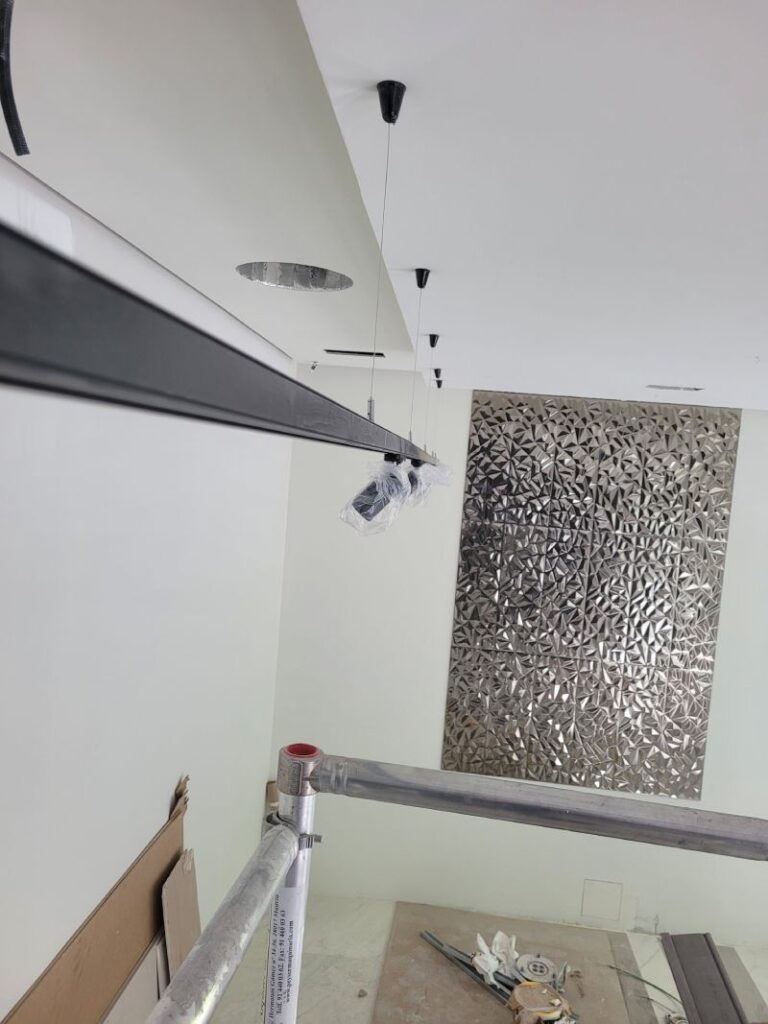 Habitación en renovación con rieles de focos LED suspendidos en el techo. Hay un andamio y materiales de construcción en el suelo. La pared tiene un panel decorativo metálico.