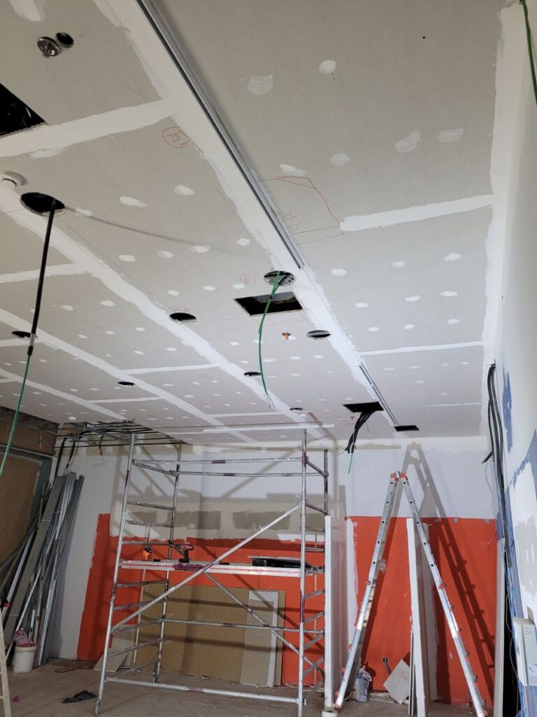 Habitación en proceso de construcción o renovación, con el techo abierto mostrando paneles de yeso parcialmente instalados y cables eléctricos colgando. Hay un andamio y una escalera en la habitación, así como varios materiales de construcción y herramientas. Las paredes están parcialmente pintadas de blanco y naranja, y se observan áreas aún sin terminar.