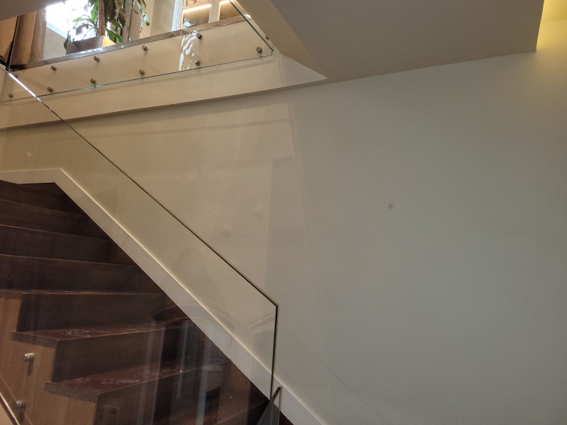 Escalera de madera con barandilla de vidrio en el interior de un edificio. La escalera sube hacia un entrepiso que tiene una barandilla de vidrio con soportes metálicos. En el entrepiso se puede ver una planta decorativa y parte de un área de estar con estanterías. Las paredes son de color claro, y el diseño es moderno y elegante.