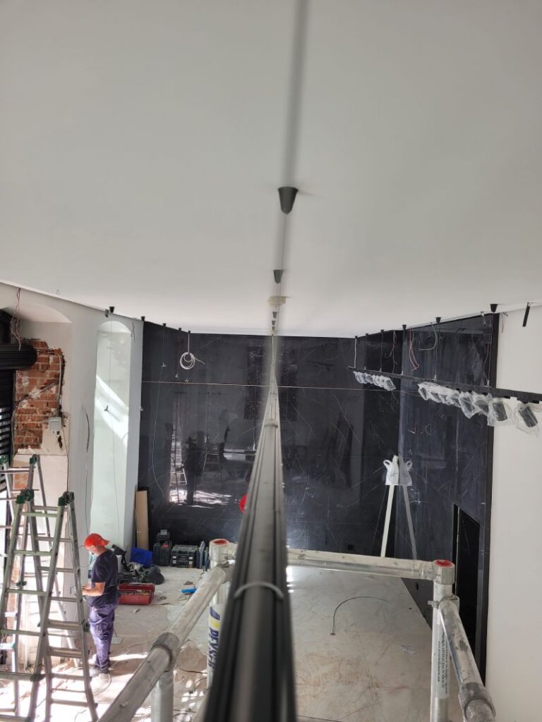 Vista desde una perspectiva elevada de un espacio en renovación con rieles de focos LED instalados en el techo. Dos trabajadores con cascos rojos y mascarillas están de pie en el lado izquierdo, cerca de una escalera y varias herramientas. Al fondo, se observa una pared de mármol negro y una escalera de tijera. El área está llena de equipos y materiales de construcción, y el techo es blanco y liso.