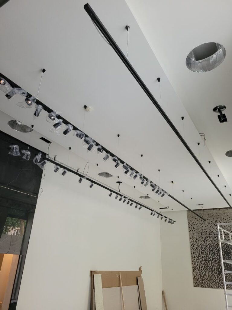 Rieles con múltiples focos LED instalados en el techo de una habitación en proceso de instalación o renovación. Los focos están parcialmente cubiertos con plástico protector. En la parte inferior se observan materiales de construcción, como paneles de yeso y cubetas. La pared de la derecha es blanca y lisa, mientras que a la izquierda hay una estructura de mármol negro y una puerta abierta.