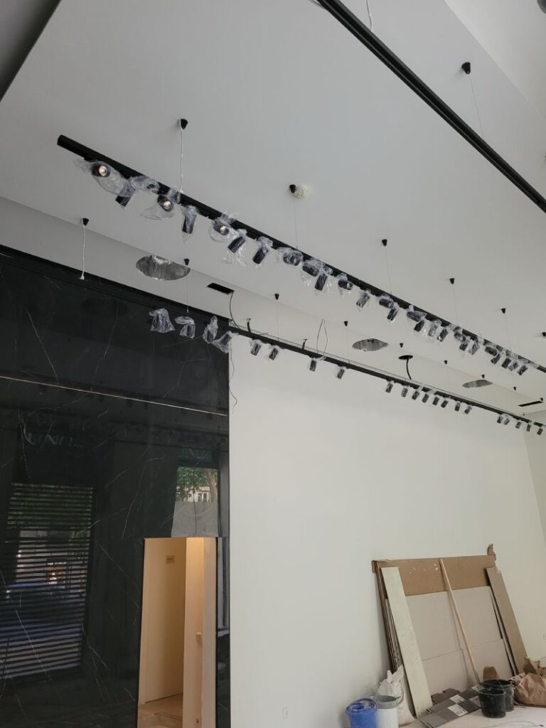 Rieles con múltiples focos LED instalados en el techo de una habitación en proceso de instalación o renovación. Los focos están parcialmente cubiertos con plástico protector. En la parte inferior se observan materiales de construcción, como paneles de yeso y cubetas. La pared de la derecha es blanca y lisa, mientras que a la izquierda hay una estructura de mármol negro y una puerta abierta.