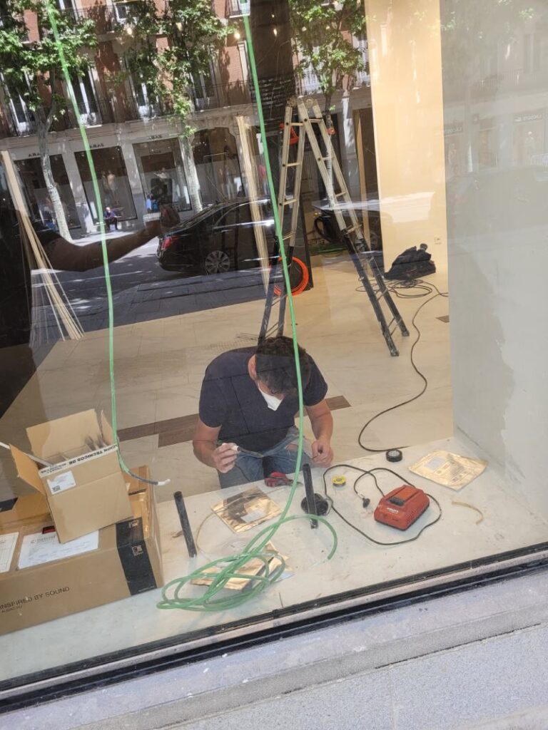 Trabajador realizando tareas de instalación eléctrica dentro de un local comercial, visto a través de la vitrina. El hombre está agachado en el suelo, manipulando cables y herramientas. Al fondo se ve una escalera, cajas de materiales y una vista de la calle con edificios y árboles reflejados en el vidrio.