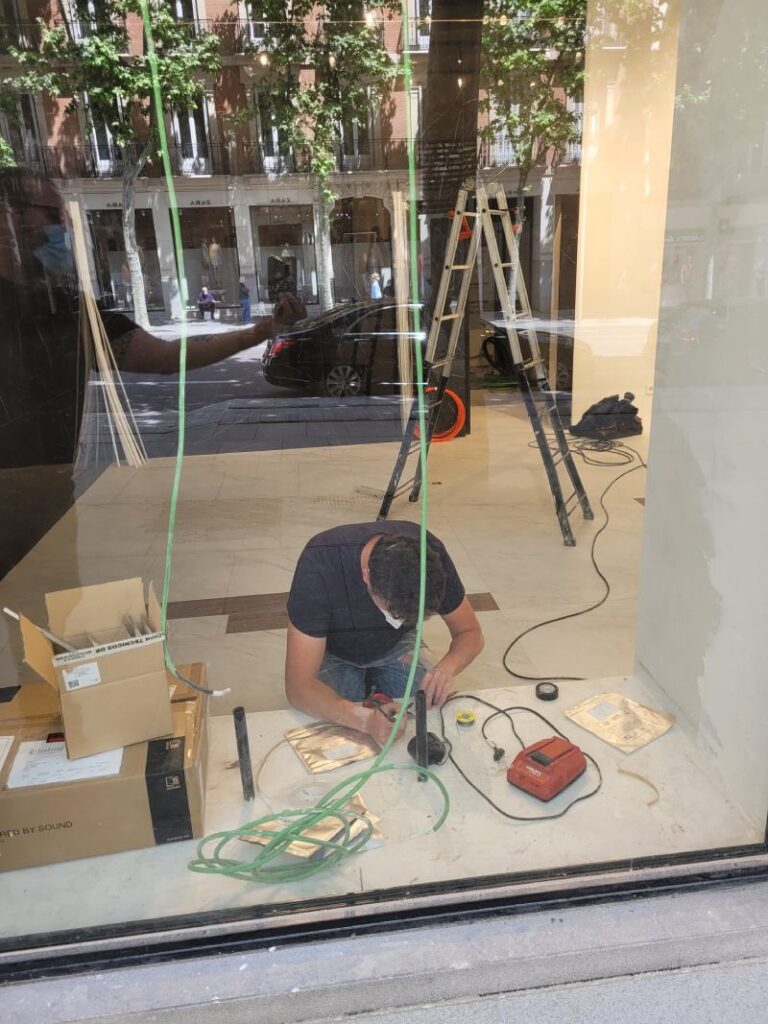 Trabajador realizando tareas de instalación eléctrica dentro de un local comercial, visto a través de la vitrina. El hombre está agachado en el suelo, manipulando cables y herramientas. Al fondo se ve una escalera, cajas de materiales y una vista de la calle con edificios y árboles reflejados en el vidrio.