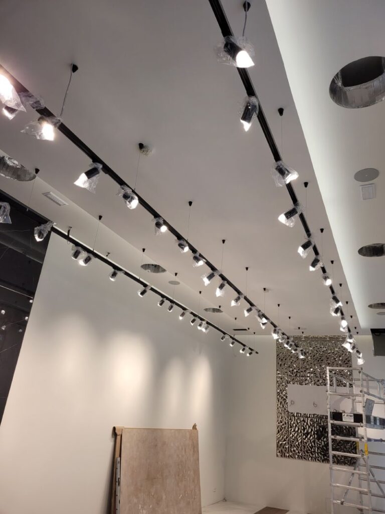 Rieles con múltiples focos LED instalados en el techo de una habitación. Los focos están parcialmente cubiertos con plástico protector. La habitación parece estar en proceso de instalación o renovación, con una escalera y materiales de construcción visibles en el fondo. La iluminación está encendida, proyectando luz sobre las paredes y el piso.