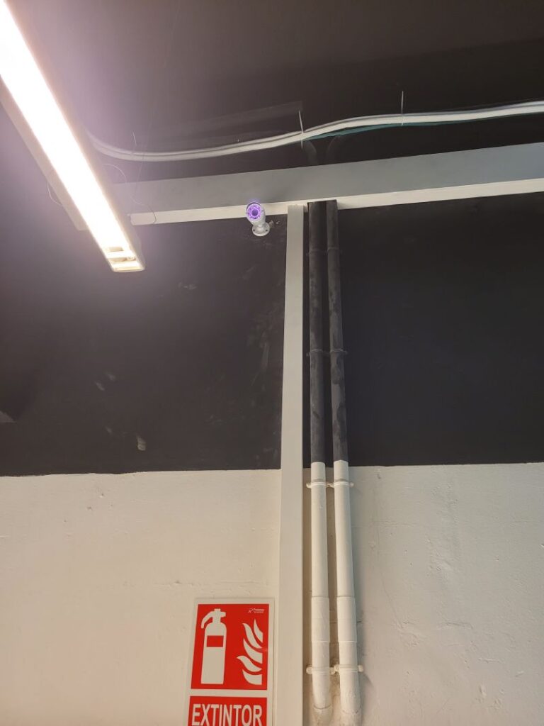 Cables organizados en canaletas en una pared blanca y negra. Letrero de extintor de incendios en la pared. Tubos verticales y una luz fluorescente también son visibles.