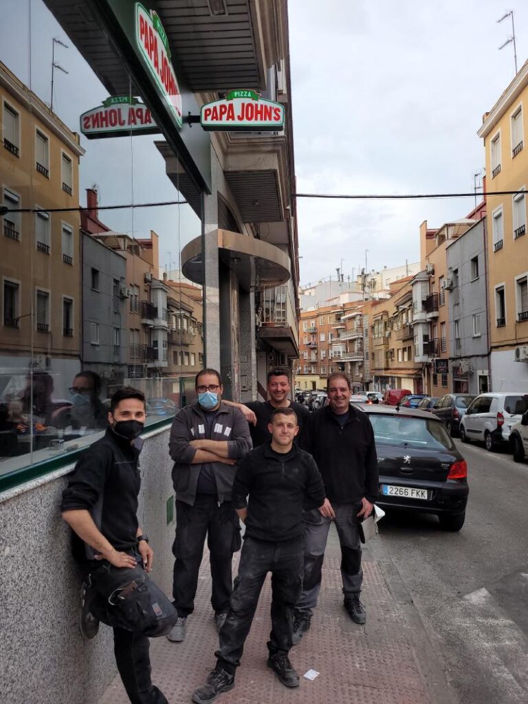 Cinco hombres vestidos con ropa de trabajo, algunos de ellos con mascarillas, posan afuera de un restaurante de Papa John's en una calle urbana. Están de pie y parecen relajados, con edificios y autos aparcados en el fondo.