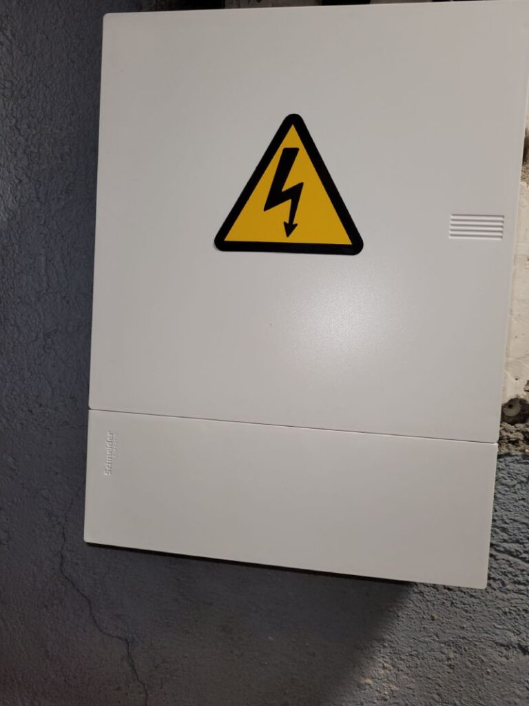 Caja de distribución eléctrica con un signo de advertencia de alto voltaje. El símbolo consiste en un triángulo amarillo con un borde negro y un rayo negro en el centro, indicando peligro eléctrico. La caja es de color blanco y parece estar montada en una pared.