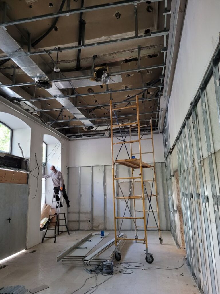 El techo está abierto, revelando la estructura, los conductos y el cableado eléctrico. Hay un andamio en el medio de la habitación y un hombre en una escalera trabajando cerca de una ventana en el lado izquierdo. Las paredes parecen estar parcialmente cubiertas con paneles de yeso, y hay materiales y herramientas esparcidos por el suelo.