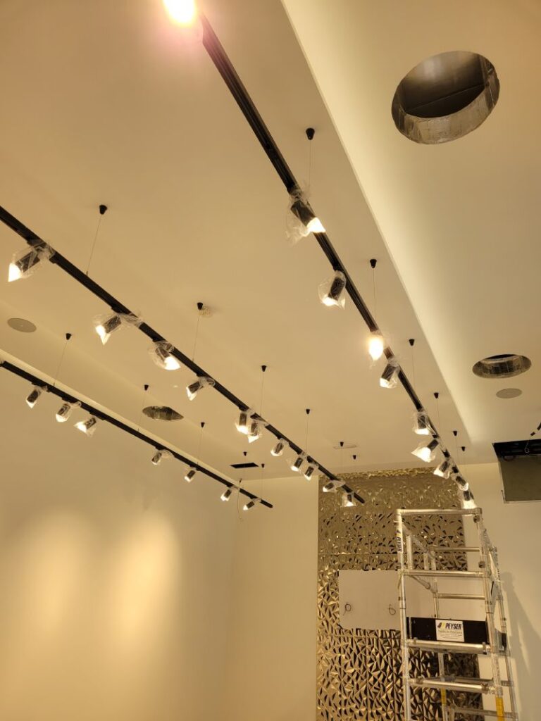 Techo de una habitación con una línea de focos colgantes y una instalación de ventilación. Las luces están empaquetadas y listas para ser instaladas, y el techo tiene un diseño moderno y limpio."