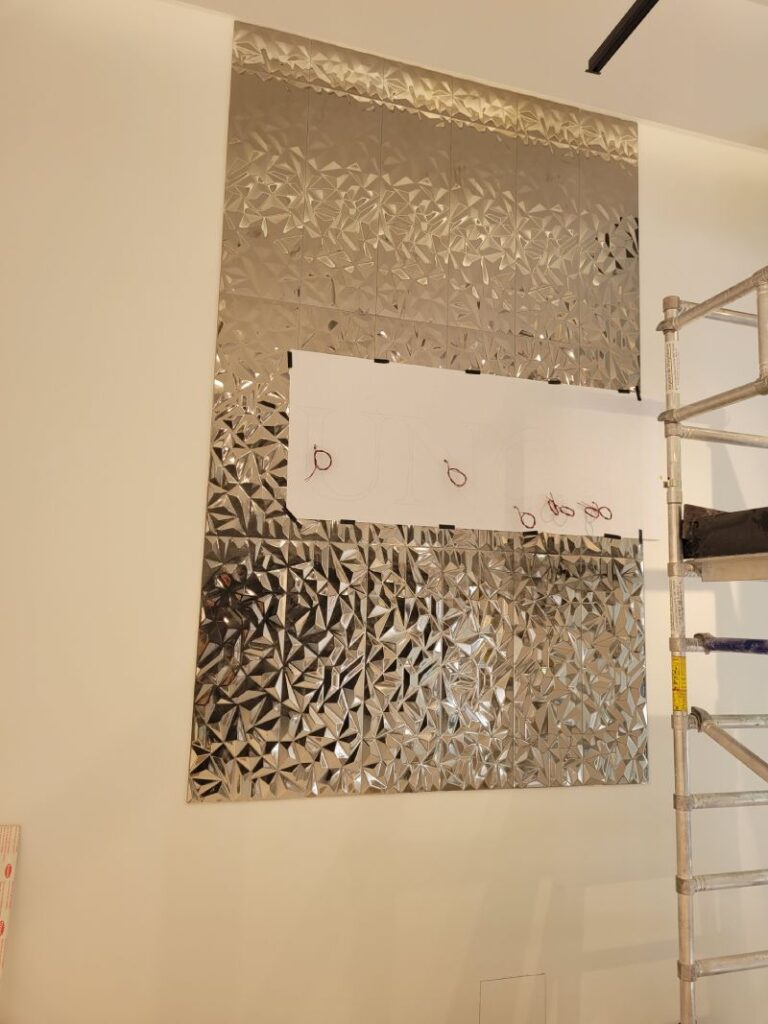 Interior de una habitación con una gran pared decorativa de metal con un diseño geométrico. Hay un andamio al lado de la pared, indicando que la habitación está en proceso de instalación o renovación.