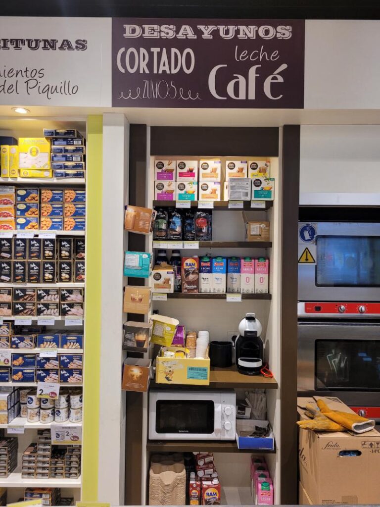 Sección de una tienda que muestra productos para el desayuno, con un letrero que dice 'Desayunos', 'Cortado', 'Leche', 'Café', 'Zumos'. Hay estantes llenos de cajas de café y otros productos relacionados con el desayuno, junto con un microondas y una cafetera en la parte inferior.