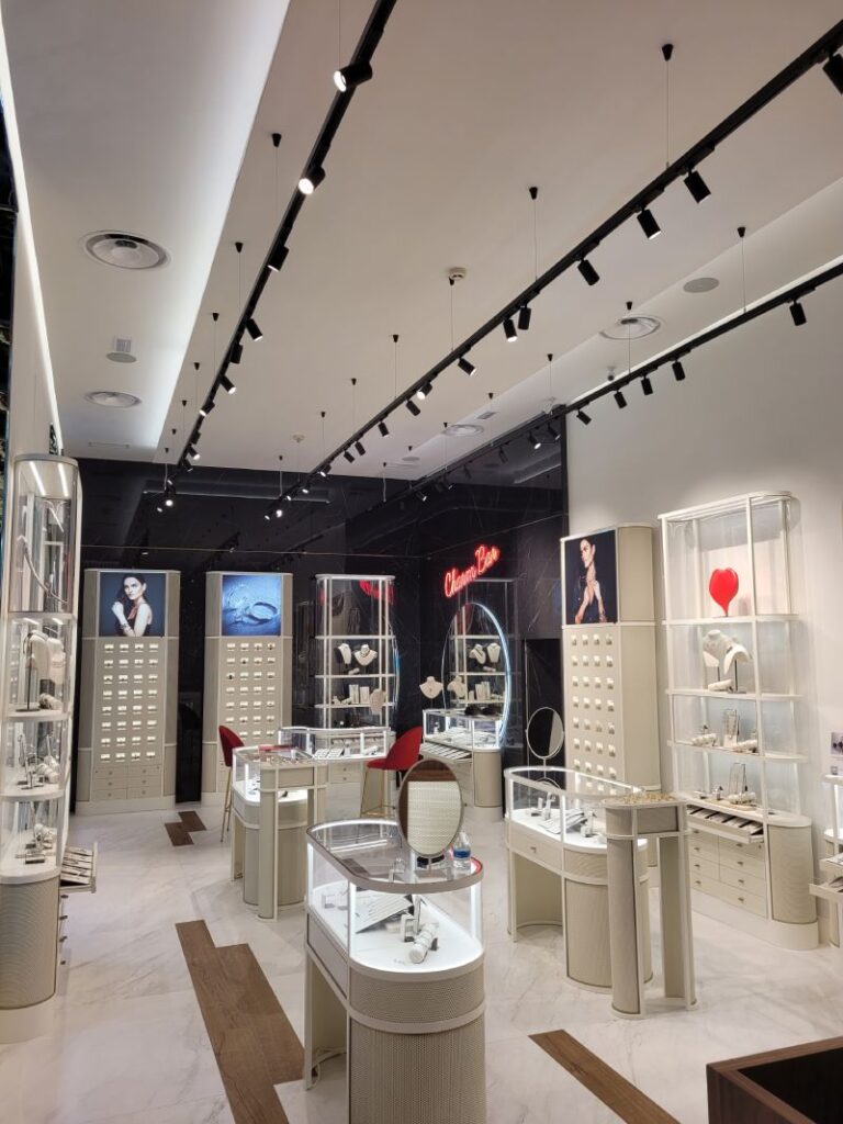 Tienda de joyería con vitrinas elegantes, luces en rieles negros en el techo y decoración moderna. Expositores iluminados muestran diversas piezas de joyería. El ambiente es sofisticado, con suelos de mármol y sillas rojas para atención al cliente.