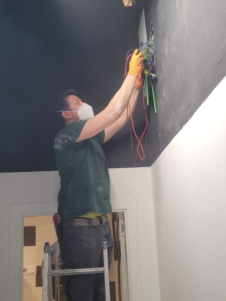 Técnico trabajando en una pared, realizando conexiones eléctricas. El técnico está en una escalera, lleva una mascarilla y guantes naranjas. La pared es de color oscuro y la habitación parece estar en proceso de construcción o renovación.
