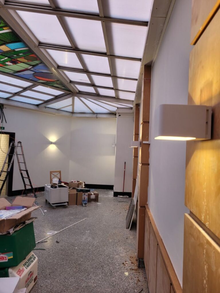 Interior de una habitación con techo de vidrio y paredes de color claro. Se observan varias cajas y materiales de construcción en el suelo, una escalera en la esquina y apliques de luz en las paredes.