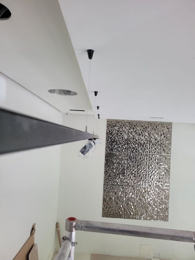Interior de una habitación en construcción con un sistema de iluminación colgante recién instalado. Hay una estructura metálica en el fondo con un diseño geométrico y materiales de construcción esparcidos por el suelo.