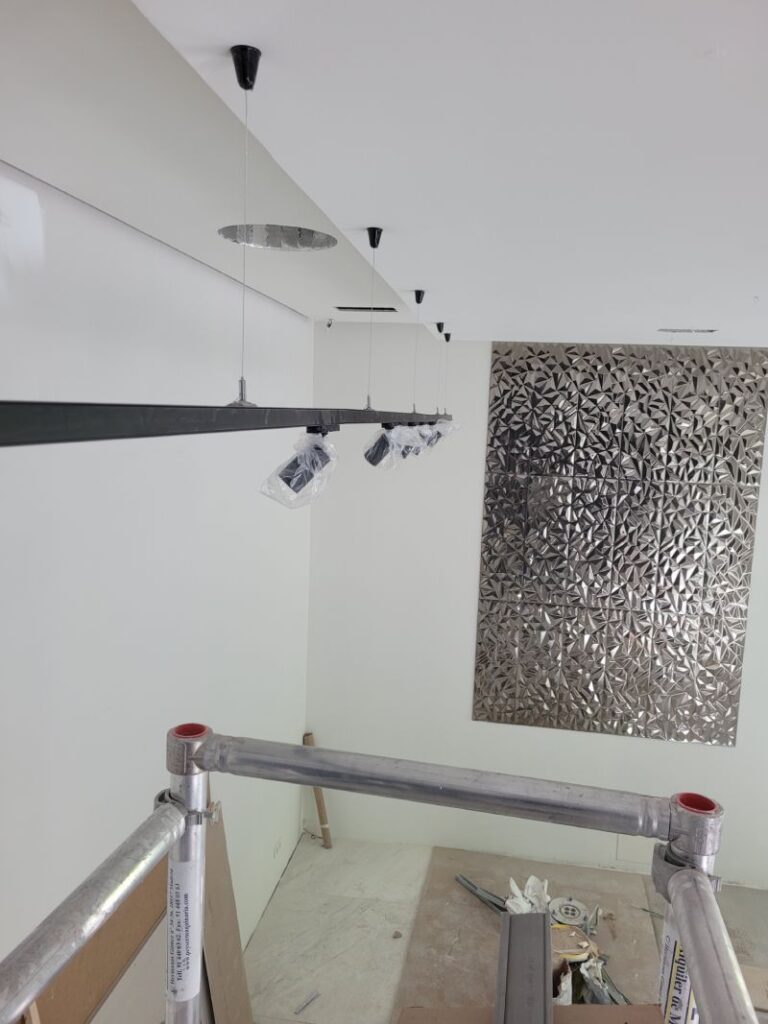 Interior de una habitación en construcción con un sistema de iluminación colgante recién instalado. Hay una estructura metálica en el fondo con un diseño geométrico y materiales de construcción esparcidos por el suelo.