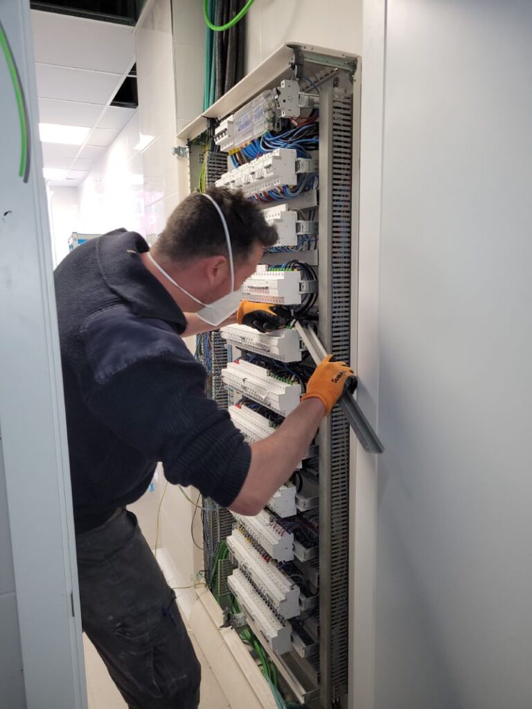 Técnico trabajando en un gabinete eléctrico abierto, ajustando componentes dentro del gabinete. El técnico lleva una mascarilla y guantes naranjas. El gabinete contiene múltiples disyuntores y cables organizados.