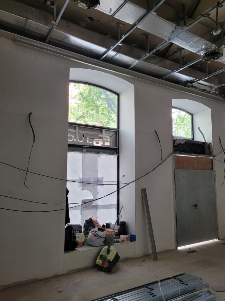 Interior de una habitación en construcción con dos ventanas grandes y un techo abierto que muestra conductos y estructura metálica. Hay materiales de construcción y herramientas esparcidos por la habitación.