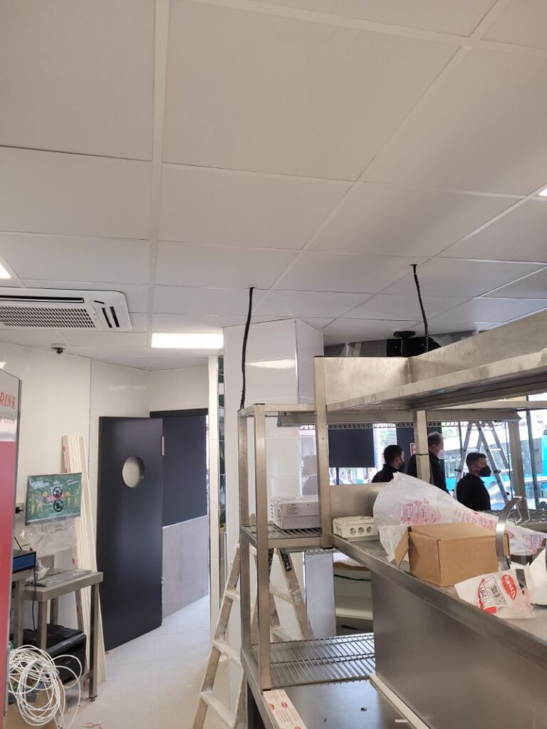 Interior de una tienda de Papa John's en construcción, con trabajadores en el fondo. Hay estanterías metálicas, escaleras de mano y herramientas dispersas en el área de trabajo. El techo está parcialmente instalado con paneles de iluminación.