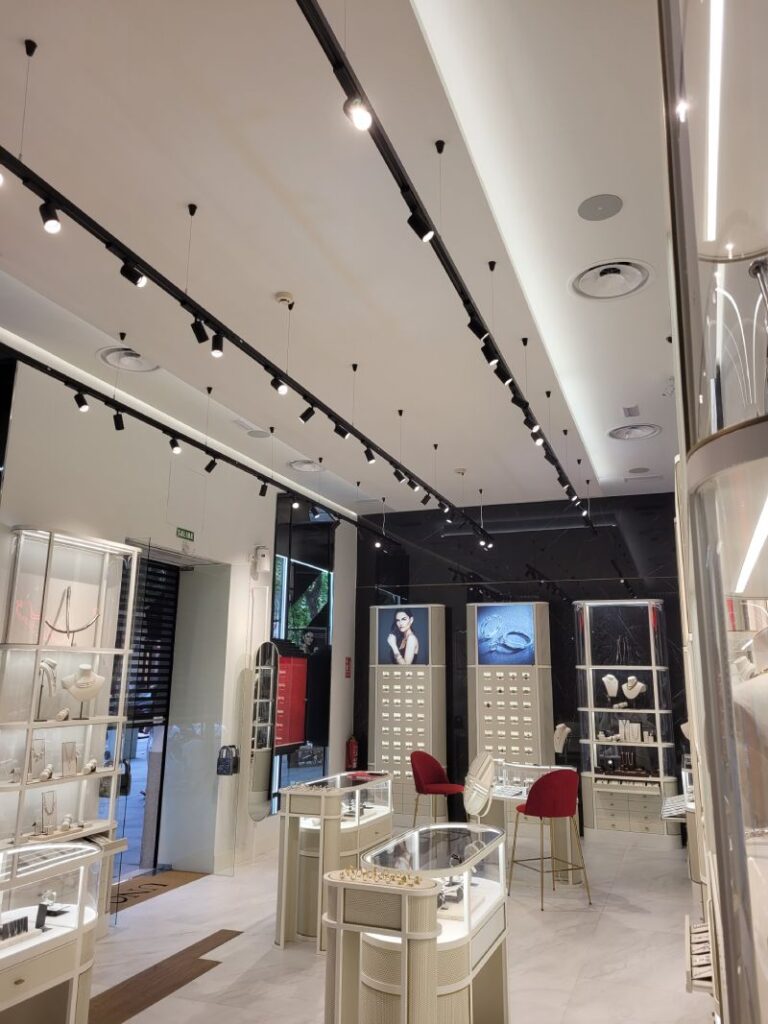 Tienda de joyería con vitrinas elegantes, luces en rieles negros en el techo y decoración moderna. Expositores iluminados muestran diversas piezas de joyería. El ambiente es sofisticado, con suelos de mármol y sillas rojas para atención al cliente.