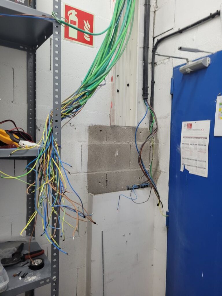 Área técnica con cables eléctricos desordenados colgando de la pared, estante metálico con herramientas y puerta azul con documentos. Señal de extintor en la pared.