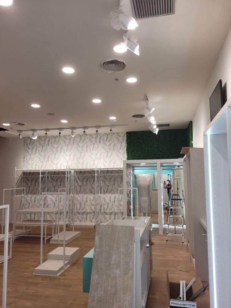 Tienda en montaje con estanterías blancas, papel pintado de hojas y pared con follaje artificial. Suelo de madera, focos en el techo y estructuras para exhibir productos. Hay una escalera y cajas de materiales en el fondo.