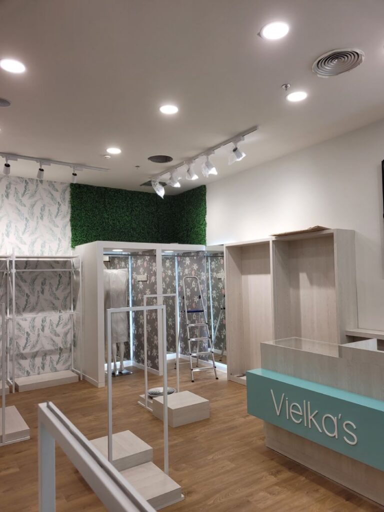 Tienda en montaje con estanterías blancas y mostrador con el nombre "Vielka's". Pared decorada con papel pintado de hojas y secciones de follaje artificial. Suelo de madera, lámparas colgantes y estructura metálica para exhibir productos.