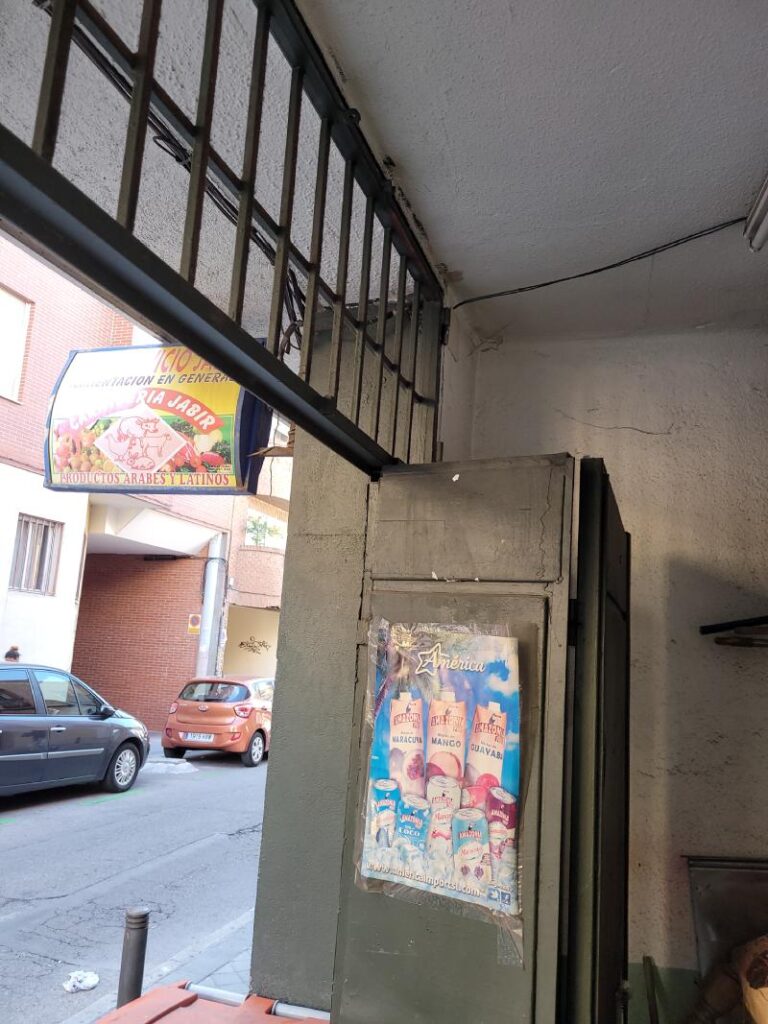 Entrada de una tienda de alimentación con una reja en el techo y un cartel publicitario. En el exterior se pueden ver varios coches aparcados en la calle.
