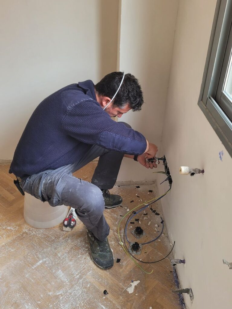 Hombre trabajando en la instalación de cables eléctricos en una pared, sentado en un taburete improvisado con una mascarilla bajada. Alrededor hay cables sueltos y herramientas en el suelo de parquet.