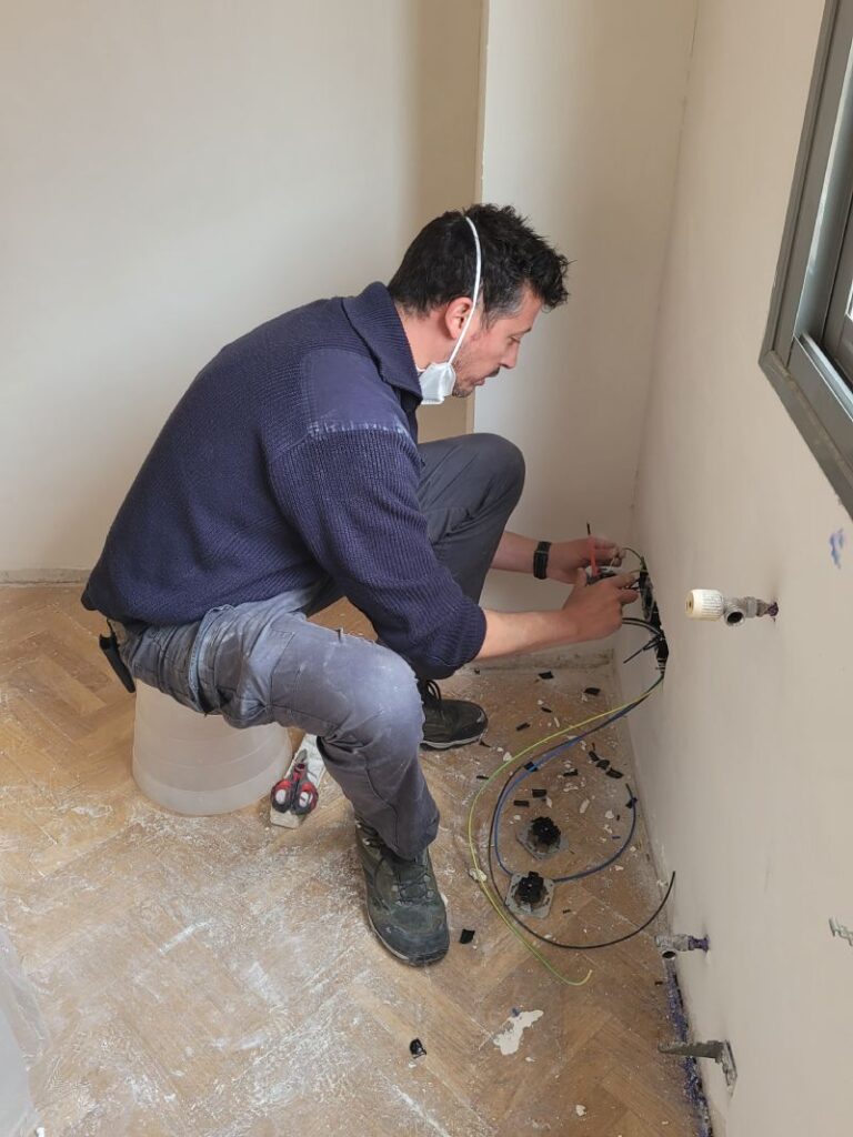 Hombre trabajando en la instalación de cables eléctricos en una pared, sentado en un taburete improvisado con una mascarilla bajada. Alrededor hay cables sueltos y herramientas en el suelo de parquet.