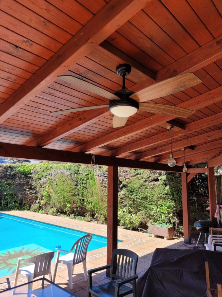 Vista de un patio cubierto con techo de madera, mostrando un ventilador de techo y luces colgantes. En el fondo hay una piscina azul rodeada de sillas y vegetación. El ambiente es relajado y apropiado para actividades al aire libre.