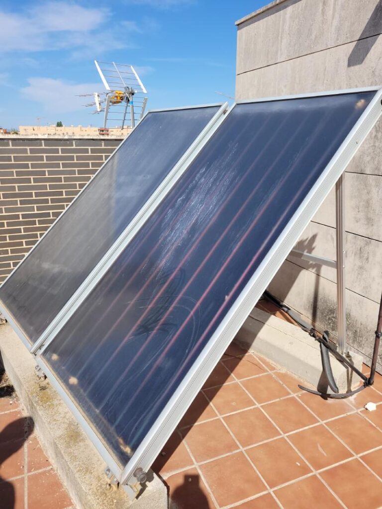 Paneles solares instalados en el techo de un edificio bajo un cielo despejado. Los paneles están inclinados para captar la luz solar y están conectados a un sistema de soporte metálico. Al fondo se ven otras construcciones y una antena.