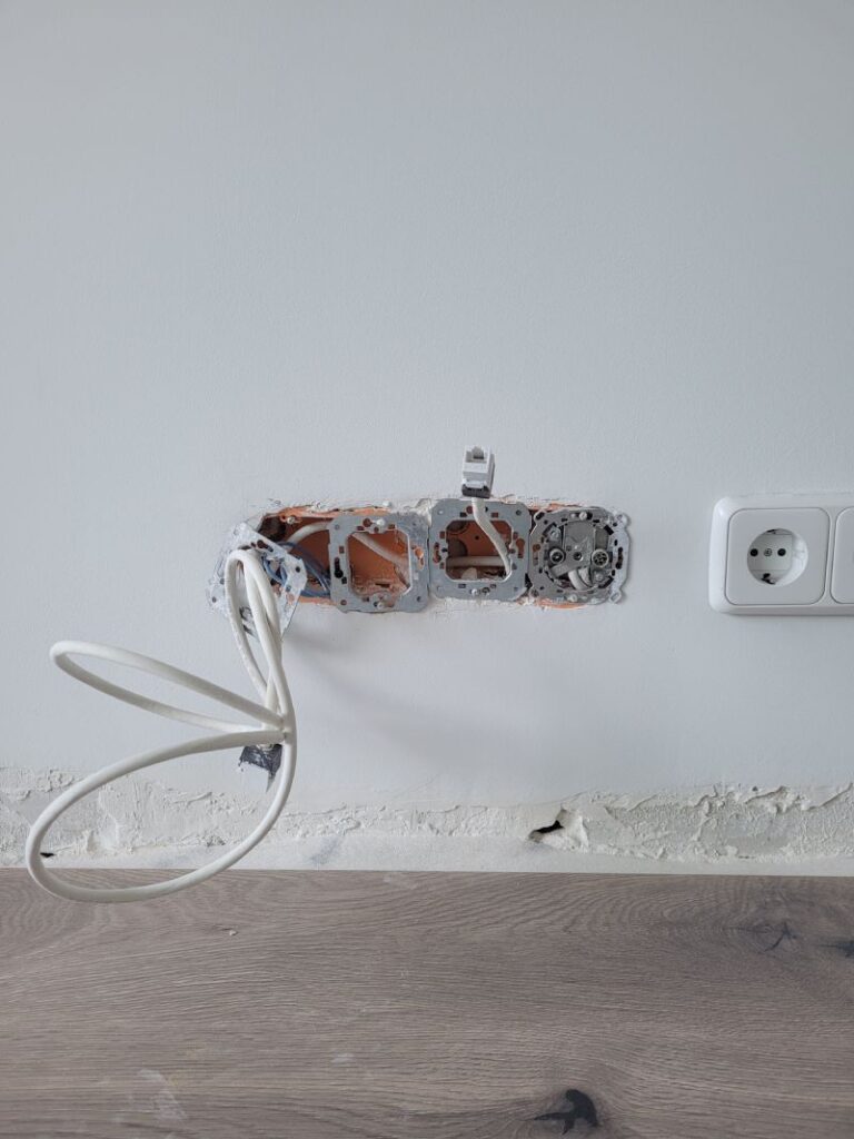 Vista de una pared con una instalación de enchufes en progreso, mostrando cajas de enchufe sin cubrir y cables sueltos. A la derecha hay un enchufe ya instalado en la pared.