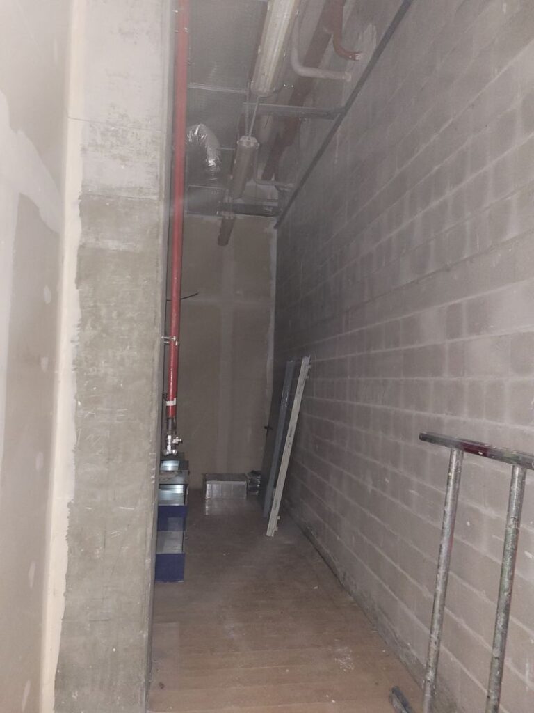 Pasillo estrecho y sin terminar con paredes de bloques de hormigón y varias tuberías expuestas en el techo. Hay algunos materiales y herramientas de construcción en el suelo.