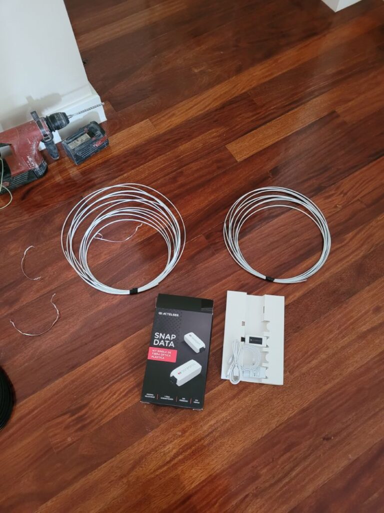 Herramientas y materiales de instalación sobre un suelo de madera, incluyendo rollos de cable blanco, un taladro rojo, y una caja etiquetada como 'Actelser SNAP DATA'.