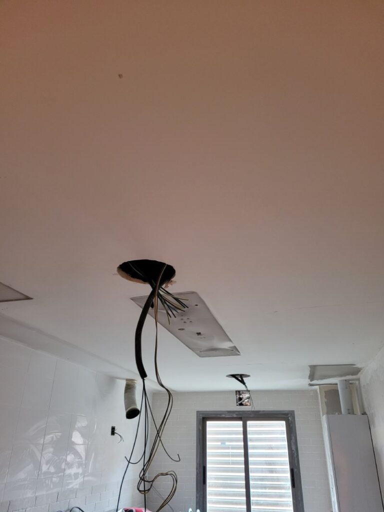 Vista de un techo con cables colgando de una abertura, en una habitación en proceso de renovación. Las paredes están alicatadas con azulejos blancos y una ventana en el fondo deja entrar la luz natural.
