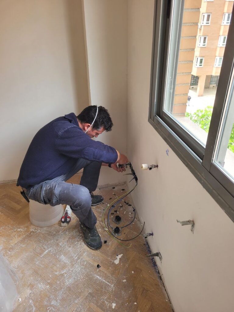 Hombre trabajando en la instalación de cables eléctricos en una pared, sentado en un taburete improvisado con una mascarilla bajada. Alrededor hay cables sueltos y herramientas en el suelo de parquet.