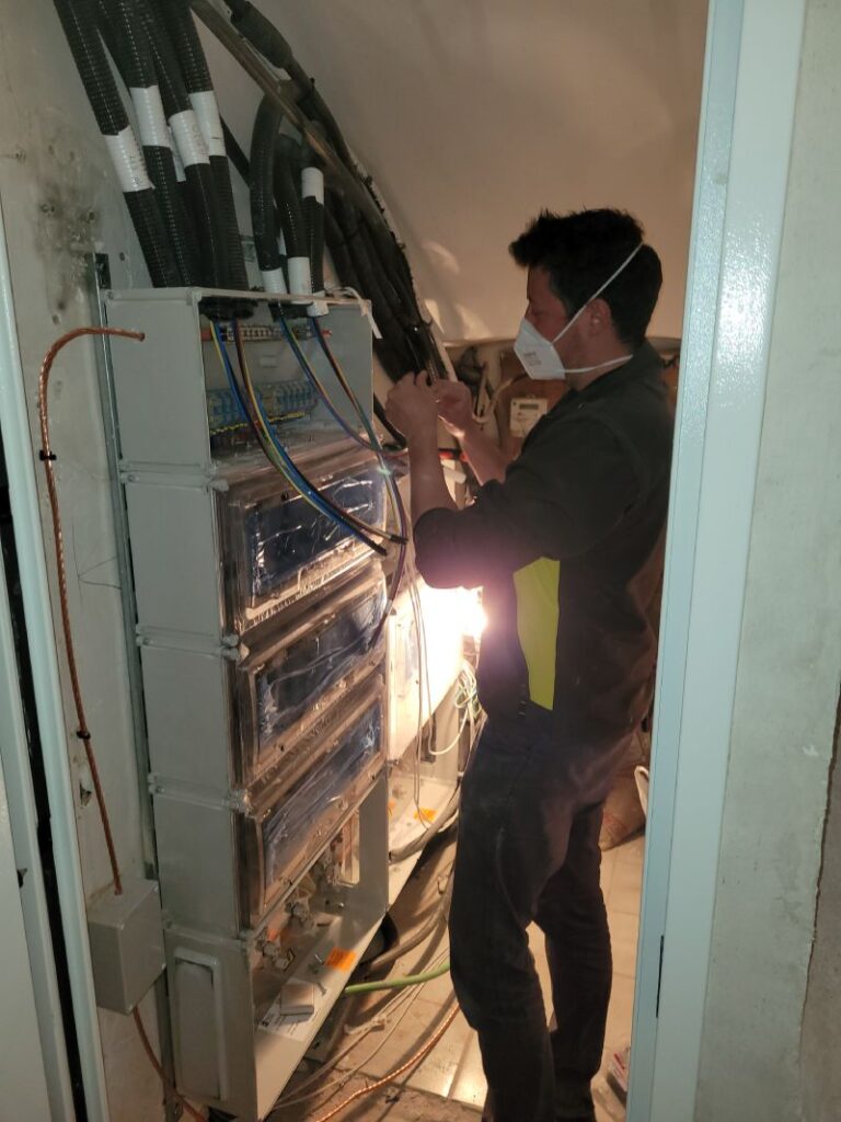 Técnico trabajando en un gabinete eléctrico abierto, ajustando componentes dentro del gabinete. El técnico lleva una mascarilla y guantes naranjas. El gabinete contiene múltiples disyuntores y cables organizados. En el fondo, se puede ver un área de trabajo con ventanas y un cartel publicitario.
