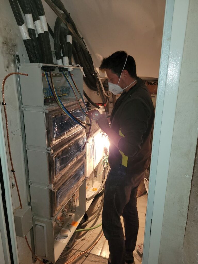 Técnico trabajando en un gabinete eléctrico abierto, ajustando componentes dentro del gabinete. El técnico lleva una mascarilla y guantes naranjas. El gabinete contiene múltiples disyuntores y cables organizados. En el fondo, se puede ver un área de trabajo con ventanas y un cartel publicitario.