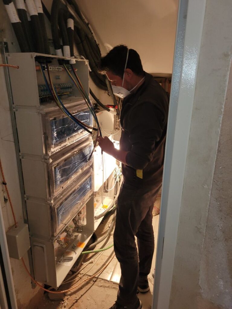 Técnico trabajando en un gabinete eléctrico abierto, ajustando componentes dentro del gabinete. El técnico lleva una mascarilla y guantes naranjas. El gabinete contiene múltiples disyuntores y cables organizados. En el fondo, se puede ver un área de trabajo con ventanas y un cartel publicitario.