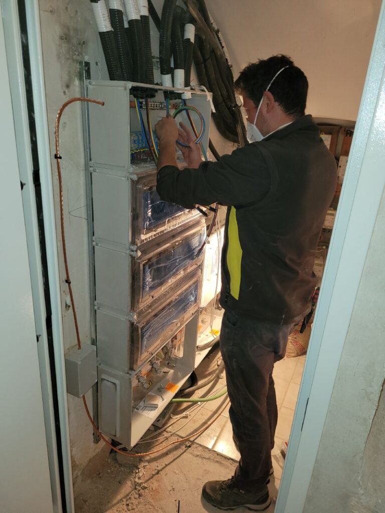 Técnico trabajando en un gabinete eléctrico abierto, ajustando componentes dentro del gabinete. El técnico lleva una mascarilla y guantes naranjas. El gabinete contiene múltiples disyuntores y cables organizados. En el fondo, se puede ver un área de trabajo con ventanas y un cartel publicitario.