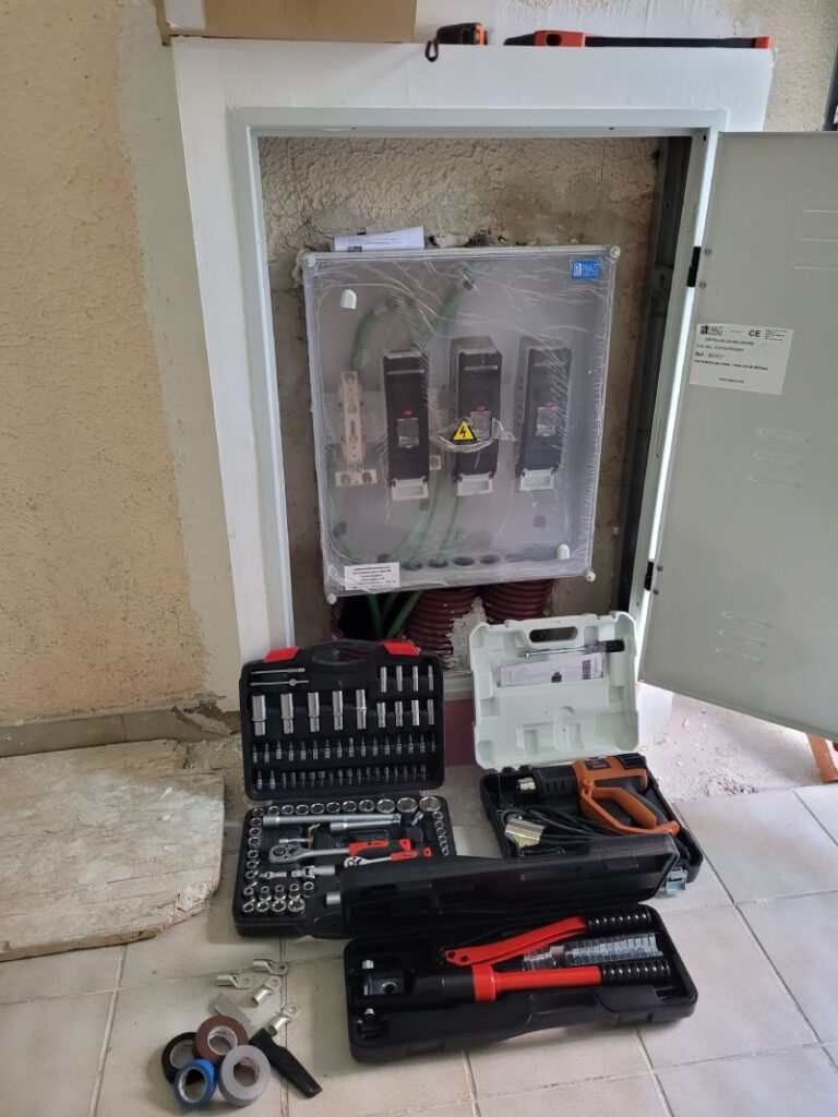 Un cuadro eléctrico abierto con cables gruesos de color verde conectados a interruptores y disyuntores. El cuadro tiene una cubierta transparente protectora. Delante del cuadro, en el suelo, hay varias cajas de herramientas abiertas que contienen diferentes tipos de llaves, destornilladores y enchufes. También hay un cortador de cables rojo, rollos de cinta adhesiva de varios colores y algunas piezas de conexión metálicas.